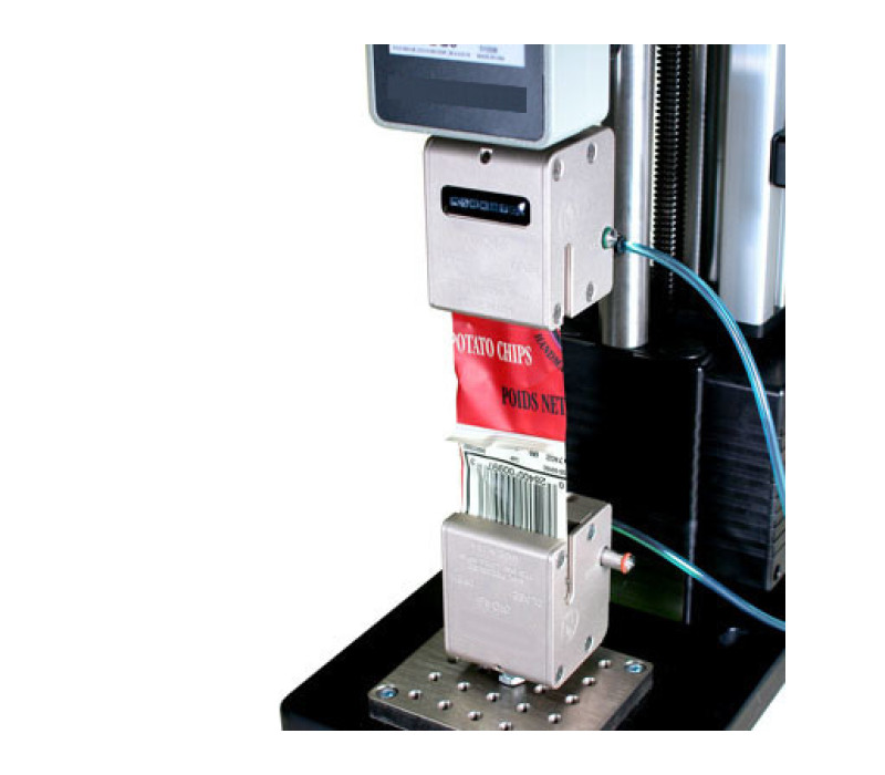 Se muestra un par de pinzas G1046 en una prueba típica de pelado a 180° de una tira de envase sellada, utilizadas junto con un dinamómetro digital y un banco de pruebas.